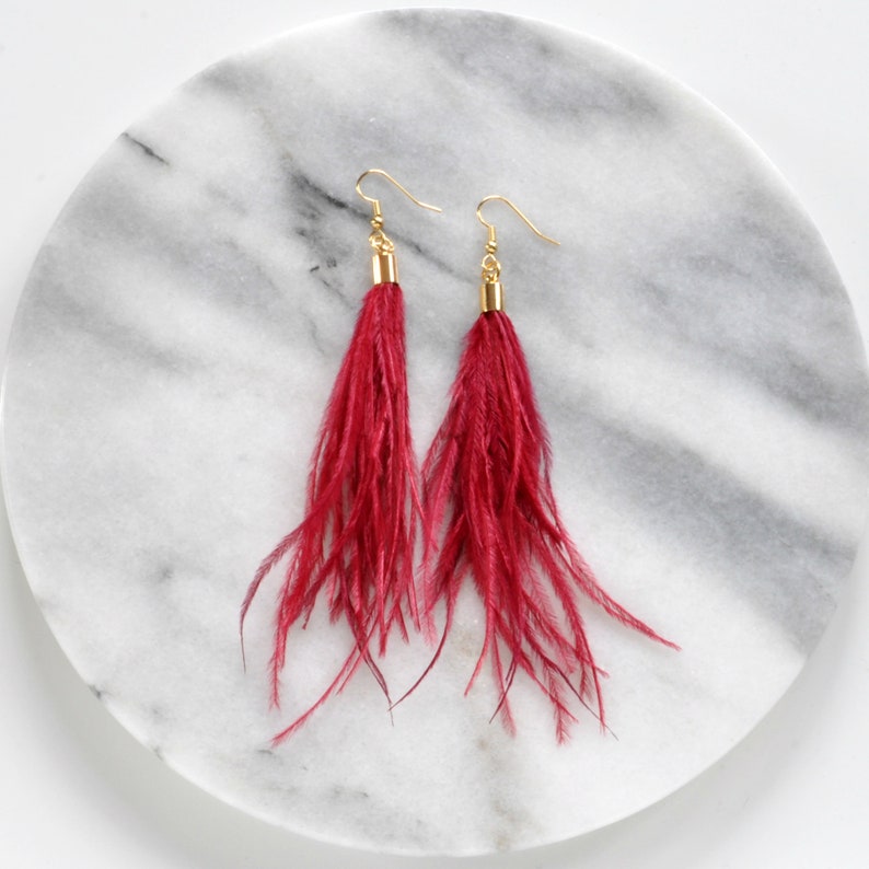May include: A pair of long, red feather earrings with gold hooks. The earrings are hanging from a white marble surface.