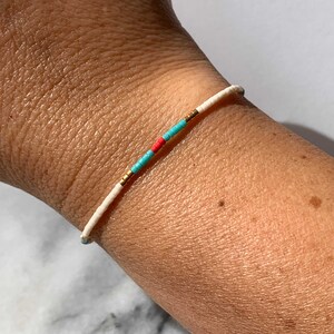 May include: A white beaded bracelet with turquoise and red accents. The bracelet is on a person's wrist.