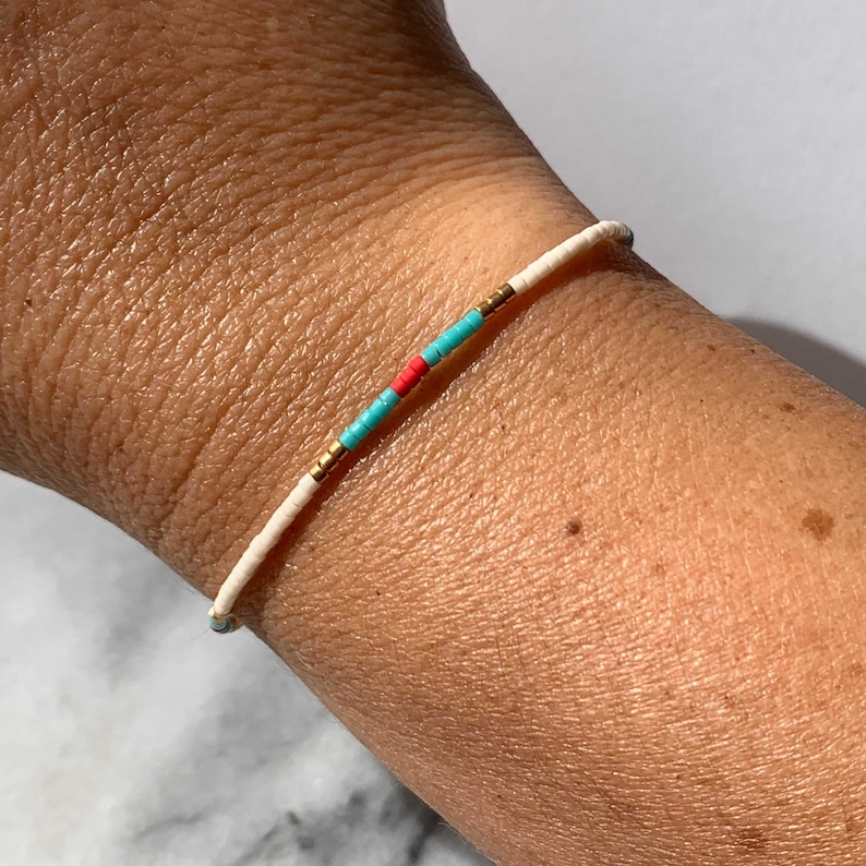May include: A white beaded bracelet with turquoise and red accents. The bracelet is on a person's wrist.