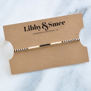 Puede incluir: Un brazalete de cuentas blancas y negras con detalles dorados. El brazalete se muestra en una tarjeta marrón con el texto "Libby & Smee Handmade in Westfield, NJ".