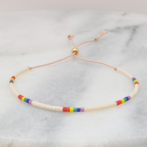 May include: A white beaded bracelet with a rainbow stripe of colored beads and a gold bead accent. The bracelet is tied with a pink cord.
