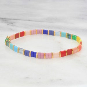 Op de afbeelding: Een kleurrijke armband met rechthoekige kralen in regenboogkleuren, waaronder rood, roze, blauw en groen. De kralen worden gescheiden door goudkleurige metalen accenten. De armband wordt getoond op een wit marmeren oppervlak.