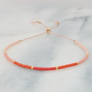 May include: A pink and orange beaded bracelet with a gold slider clasp. The bracelet is made with small, round beads and has a delicate, feminine look.