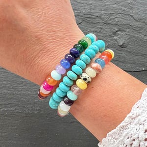 Op de afbeelding: Drie kralenarmbanden in verschillende kleuren zijn op een pols gestapeld. De armbanden hebben ronde kralen in tinten roze, oranje, turquoise, blauw, paars, groen, geel en wit. De armbanden zijn gemaakt van edelstenen.