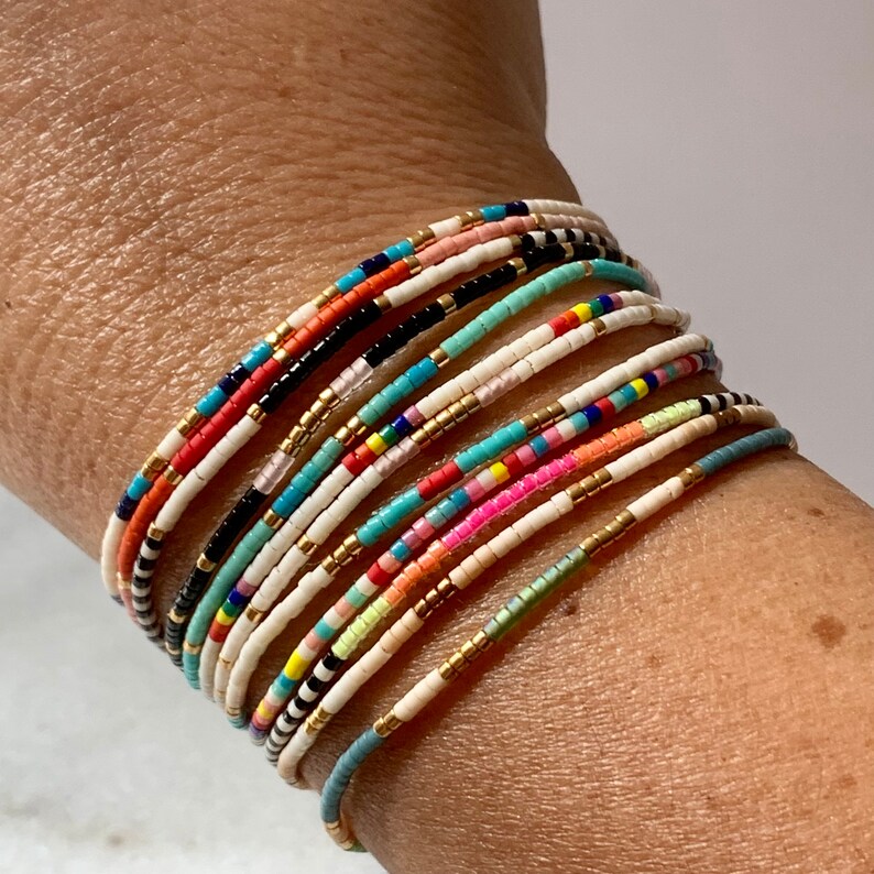 May include: A stack of colorful beaded bracelets on a person's wrist. The bracelets are made of small, round beads in various colors, including white, black, red, orange, yellow, green, blue, and pink.
