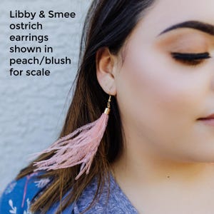 May include: A pair of blush pink ostrich feather earrings. The earrings are shown on a model for scale. Libby & Smee ostrich earrings shown in peach/blush for scale.