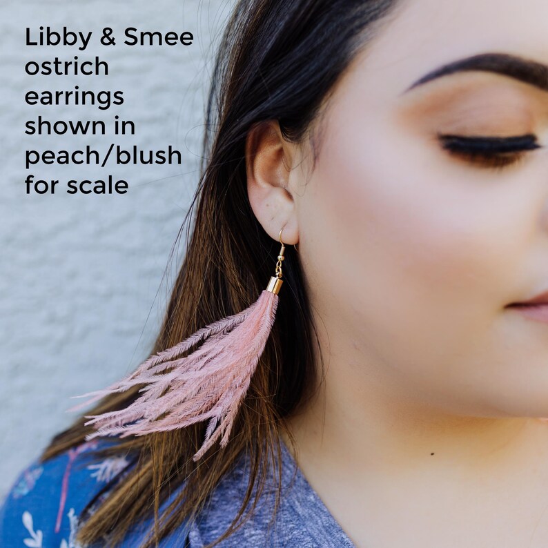 May include: A pair of blush pink ostrich feather earrings. The earrings are shown on a model for scale. Libby & Smee ostrich earrings shown in peach/blush for scale.