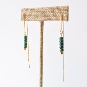 May include: A pair of gold chain earrings with green faceted beads hanging from the chains. The earrings are displayed on a brown burlap earring stand.
