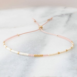 Puede incluir: Un delicado brazalete ajustable con cuentas blancas y rosadas y detalles dorados. El brazalete está hecho con un cordón rosa fino.