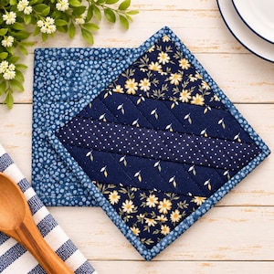 May include: A square quilted potholder featuring a diagonal patchwork design with navy blue fabric, white polka dots, and yellow daisy floral patterns. The edges are finished with a blue and white dotted fabric binding, creating a structured look.