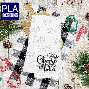 May include: A white kitchen towel with black text that reads "Cheese makes everything taste better". A black and white illustration of a wedge of cheese is above the text. The towel is on a white wooden surface with a black and white checkered fabric and holiday decorations.
