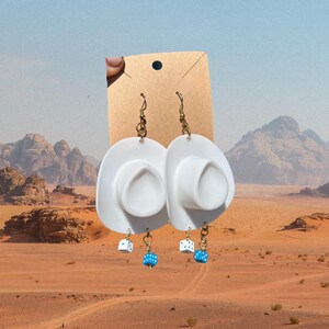 May include: White cowboy hat earrings with blue and white dice charms hanging from the bottom. The earrings are on a brown card.