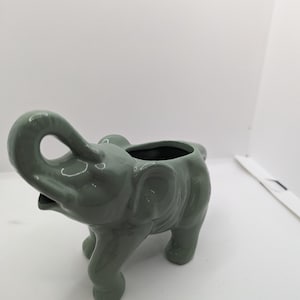 Puede incluir: Un macetero de elefante de cer&aacute;mica verde con una peque&ntilde;a abertura para plantar. El elefante tiene la trompa levantada y detalles elaborados.
