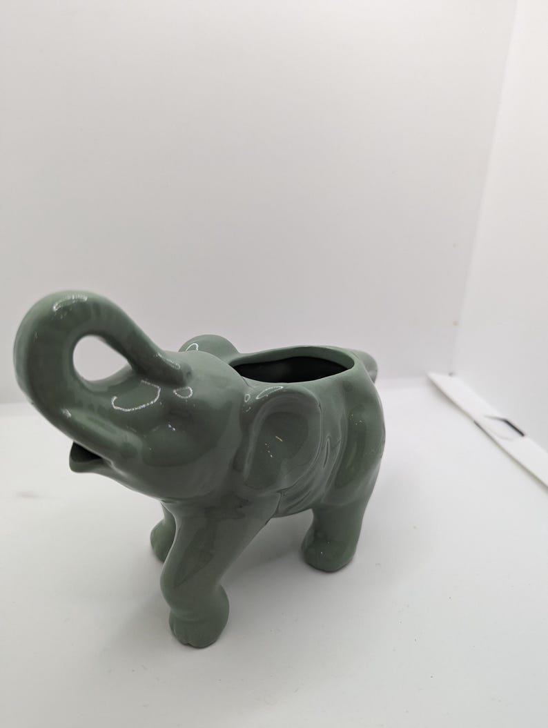 Puede incluir: Un macetero de elefante de cer&aacute;mica verde con una peque&ntilde;a abertura para plantar. El elefante tiene la trompa levantada y detalles elaborados.