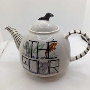 May include: White ceramic teapot with a black and white striped handle and spout. The teapot has a black bird finial on the lid and features the words "The Pour" with animal illustrations.