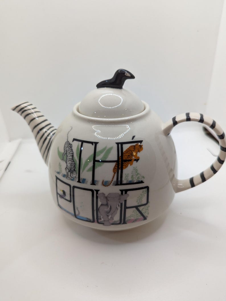 May include: White ceramic teapot with a black and white striped handle and spout. The teapot has a black bird finial on the lid and features the words "The Pour" with animal illustrations.