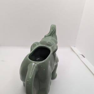 Puede incluir: Un creamer de cer&aacute;mica verde en forma de elefante con la trompa levantada y un acabado liso y esmaltado.
