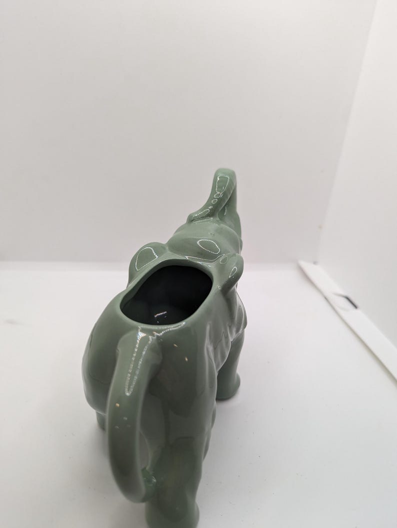 Puede incluir: Un creamer de cer&aacute;mica verde en forma de elefante con la trompa levantada y un acabado liso y esmaltado.