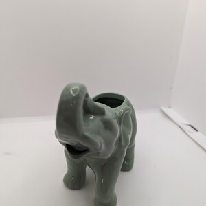 Puede incluir: Un macetero de elefante de cer&aacute;mica verde con una superficie texturizada. El macetero tiene una abertura redonda en la parte superior para plantar.
