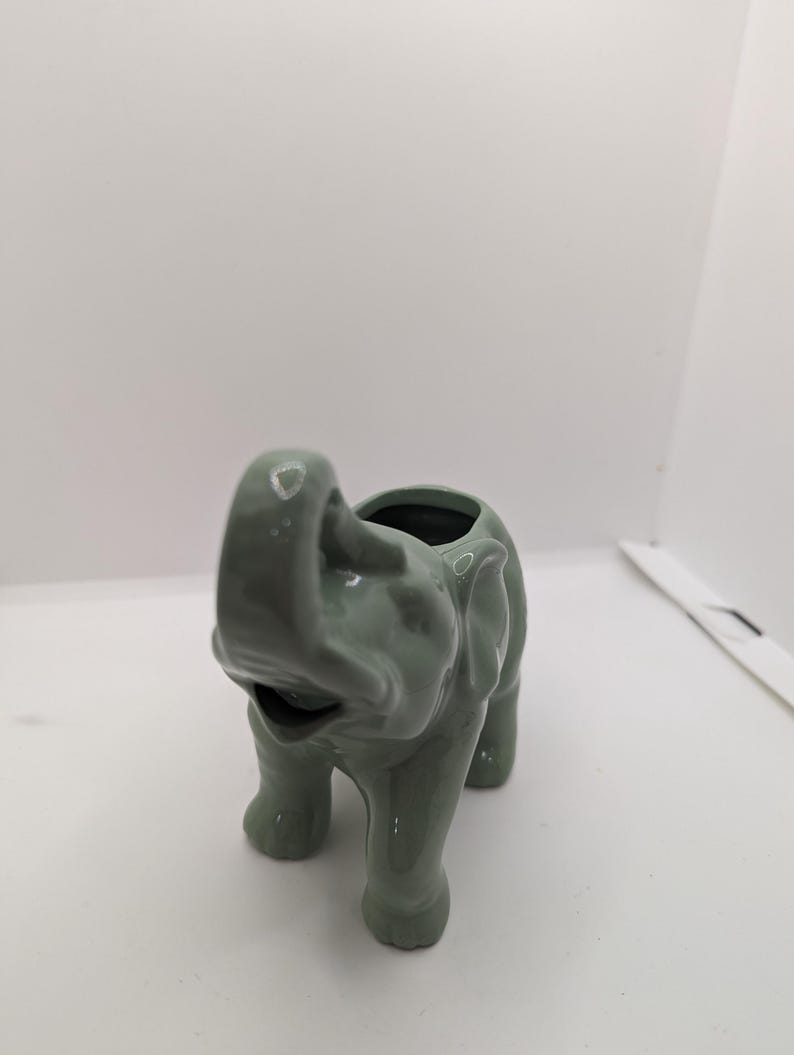 Puede incluir: Un macetero de elefante de cer&aacute;mica verde con una superficie texturizada. El macetero tiene una abertura redonda en la parte superior para plantar.