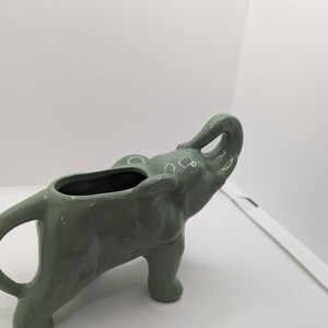 Puede incluir: Un macetero de cer&aacute;mica verde en forma de elefante con la trompa levantada. El macetero est&aacute; dise&ntilde;ado para contener plantas o flores peque&ntilde;as.