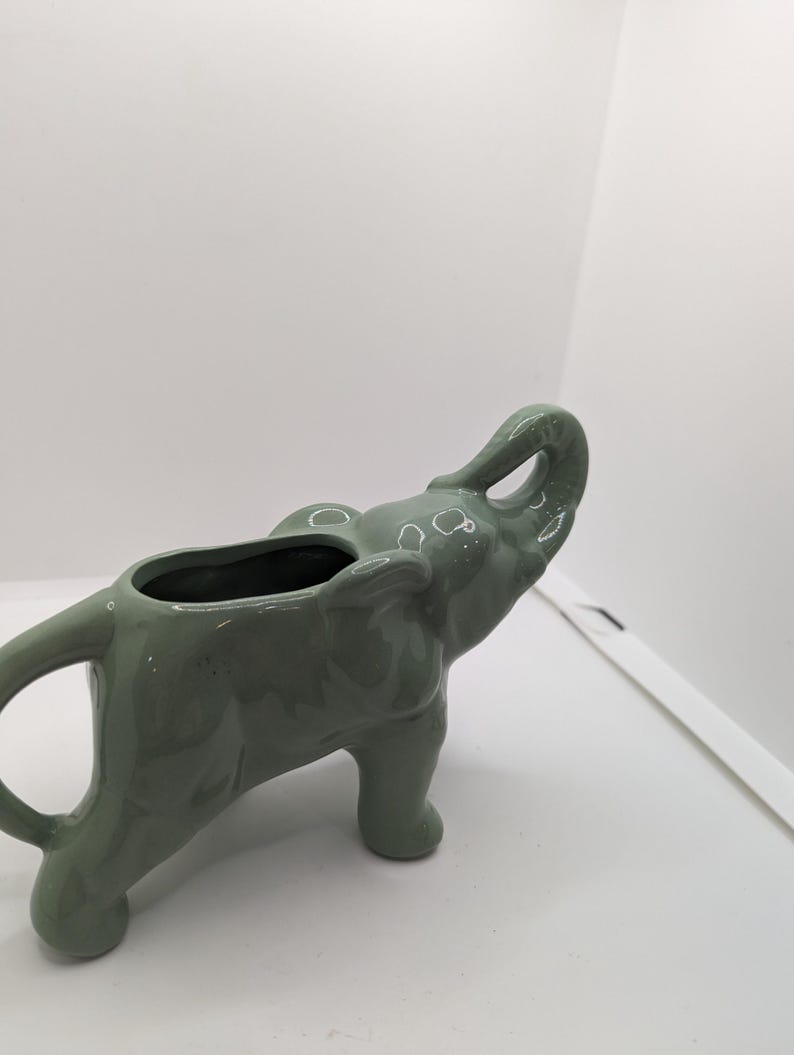 Puede incluir: Un macetero de cer&aacute;mica verde en forma de elefante con la trompa levantada. El macetero est&aacute; dise&ntilde;ado para contener plantas o flores peque&ntilde;as.