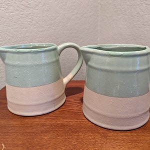 May include: Two ceramic pitchers with a light blue top and a white bottom. The pitchers have a rounded shape and a handle on the side.