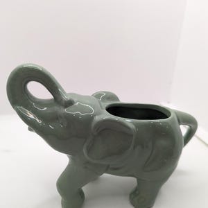 Puede incluir: Un macetero de cer&aacute;mica verde en forma de elefante con la trompa levantada y una boca ancha abierta. El macetero tiene un acabado liso y brillante.