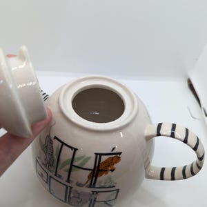 May include: White ceramic teapot with a black and white striped handle. The teapot has a lid and is decorated with a colorful illustration of animals and the word "THE" in a stylized font.
