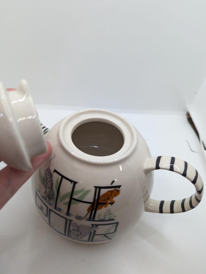 May include: White ceramic teapot with a black and white striped handle. The teapot has a lid and is decorated with a colorful illustration of animals and the word "THE" in a stylized font.