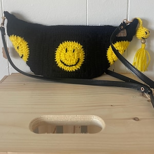 May include: Black crocheted fanny pack with a yellow smiley face design. The bag has a black adjustable strap and a yellow tassel.