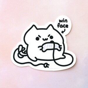 Peut inclure: Un dessin au trait noir et blanc d'un chat tenant une manette de jeu vidéo avec le texte "win face" au-dessus du chat.
