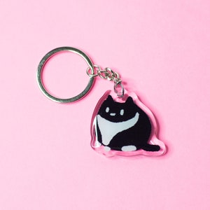 K&ouml;nnte beinhalten: Ein Schl&uuml;sselanh&auml;nger in Form einer schwarz-wei&szlig;en Katze mit einer klaren Harzbeschichtung. Der Schl&uuml;sselanh&auml;nger ist an einem silbernen Metallring befestigt.