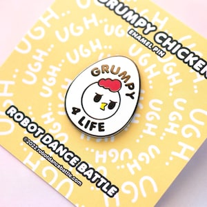 May include: White enamel pin shaped like an egg with a cartoon chicken inside. The chicken has a red comb, black eyes, and a yellow beak. The text "Grumpy 4 Life" is written in black on the egg. The pin is on a yellow background with the text "Grumpy Chicken Enamel Pin" and "Robot Dance Battle" printed on the background.