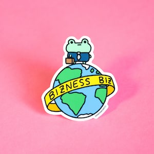 Op de afbeelding: Een sticker met een cartoonkikker in een pak die op een wereldbol staat. De wereldbol is omcirkeld door een gele banner met het woord "BIZNESS". De sticker heeft een witte rand en een roze achtergrond.