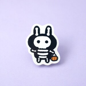 May include: A black and white cartoon bunny wearing a skeleton costume and holding a small orange pumpkin. The bunny is on a white background.