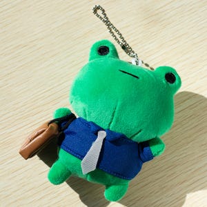 May include: Green plush frog wearing a blue suit with a gray tie and carrying a brown briefcase. The frog has black eyes and a small mouth. It is attached to a silver chain.