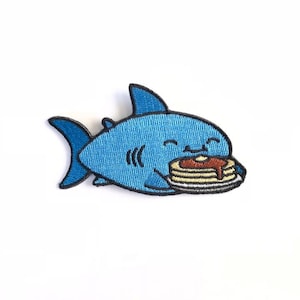 May include: A blue shark with a happy expression is holding a stack of pancakes with syrup on top. The shark is embroidered on a white background.