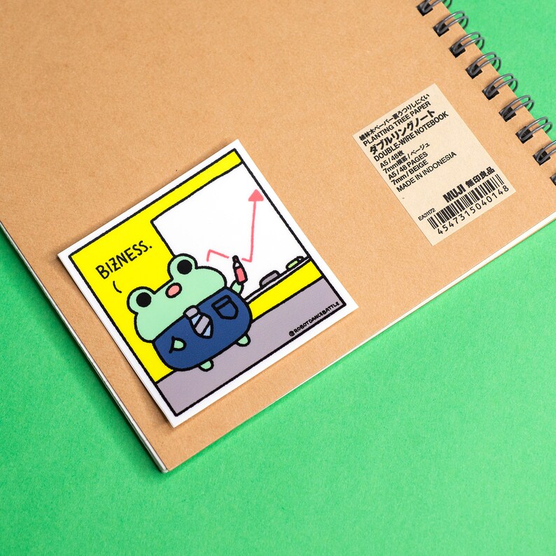 Op de afbeelding: Een gele sticker met een cartoon kikker die een blauw pak en stropdas draagt. De kikker staat voor een wit bord met een rode pijl die omhoog wijst. De tekst "BIZNESS." staat op de sticker.