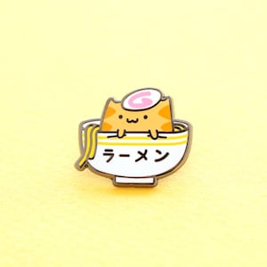 May include: A gold-colored enamel pin featuring a cartoon cat in a bowl of ramen. The cat is orange with a pink swirl on its head and the bowl says "ラーメン" in Japanese.