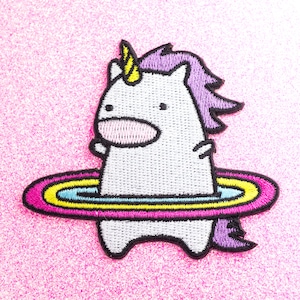Puede incluir: Un unicornio blanco con un cuerno amarillo y una crin morada está bordado sobre un fondo rosa. El unicornio está sosteniendo un aro de arcoíris rosa, amarillo y azul.