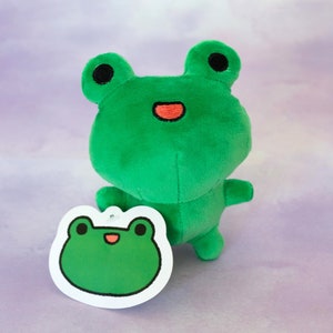 May include: Green plush frog toy with a red mouth and black eyes. The frog is standing on a white surface with a tag that has a green frog illustration on it.