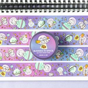 May include: A roll of glitter washi tape with a purple background and a repeating pattern of bunnies in spaceships, planets, and stars. The tape is labelled "Bunnies in Space Glitter Washi Tape 25MM x 5M".