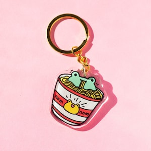 May include: A clear acrylic keychain featuring a cartoon frog eating noodles from a red and white striped bowl. The keychain has a gold metal ring and the words "nom nom" are written on the bowl.