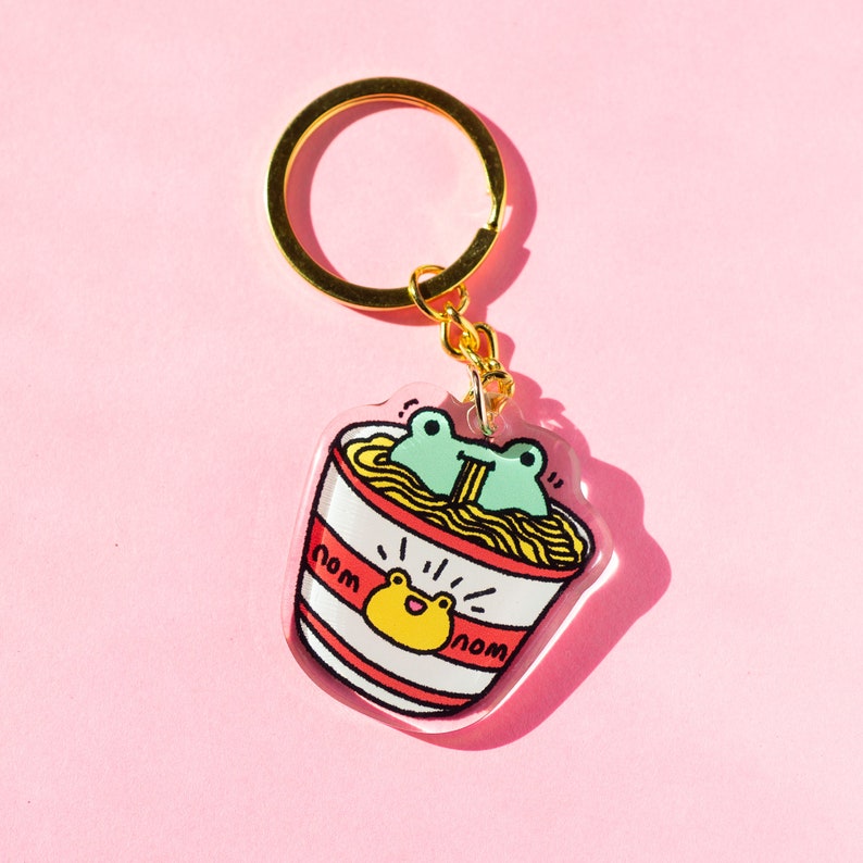 May include: A clear acrylic keychain featuring a cartoon frog eating noodles from a red and white striped bowl. The keychain has a gold metal ring and the words "nom nom" are written on the bowl.