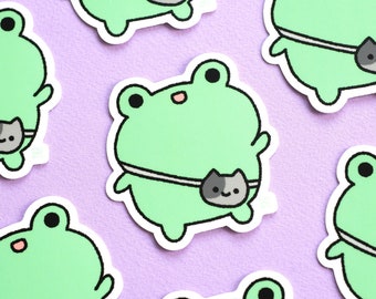 May include: A set of green frog stickers with a gray cat in a pouch on their bellies. The frogs are wearing a white strap around their bodies. The stickers are on a purple background.