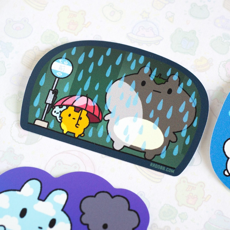 May include: A sticker featuring a cartoon illustration of two animals in the rain. The animals are a bear and a smaller animal with a red umbrella. The sticker has a green background with blue raindrops and the text "KRDBB.COM".