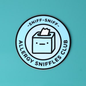 Peut inclure: Un autocollant bleu et blanc avec le texte "Allergy Sniffles Club" et "~Sniff-Sniff~" autour d'une boîte de mouchoirs de dessin animé avec un visage.