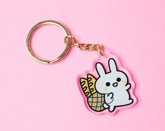 May include: A clear acrylic keychain featuring a cartoon white bunny with a basket of bread. The keychain has a gold-colored ring and chain. The background is a solid pink color.