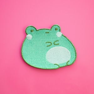 May include: A green embroidered frog patch with a white belly and white cheek spots. The frog has closed eyes and a stitched outline. The patch is on a pink background. Ideal for adding to clothing or accessories.
